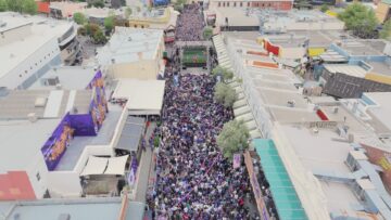 A large crowd gathers on a city street for an outdoor event, with people lining the road and a stage visible in the distance. Surrounding buildings enclose the bustling, festive atmosphere.