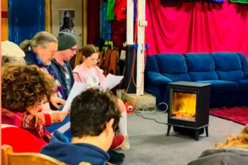 A group of people sit in a circle reading from papers near an electric space heater with a simulated flame effect.