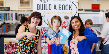 Two women and a boy stand in front of a Build a Book sign, with the boy holding a book titled Fin the Wizard.
