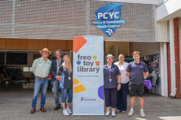 Six people stand in front of a Freo Toy Library banner and a PCYC building sign.
