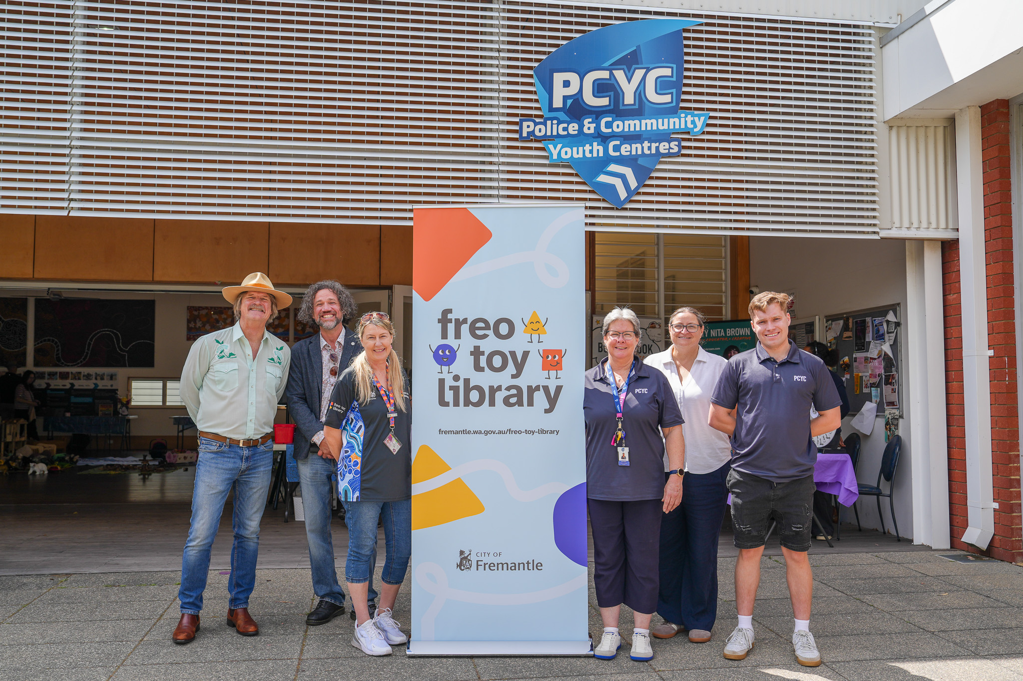 Six people stand in front of a Freo Toy Library banner and a PCYC building sign.