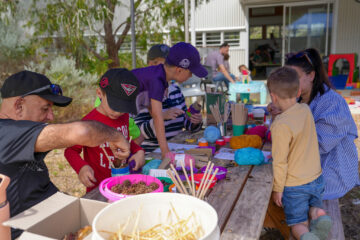 A group of children and adults sit at an outdoor table crafting with yarn, sticks, and natural materials.