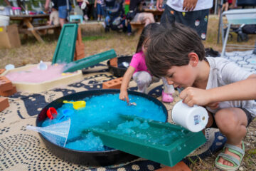 A young boy pours water from a white cup into a green tray over a large basin of blue sensory material, while a girl plays in the background.
