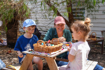 An adult and two young children sitting at a small wooden table with a basket of art supplies.