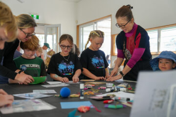 A woman in glasses assists children sitting at a table with craft supplies and paper projects.