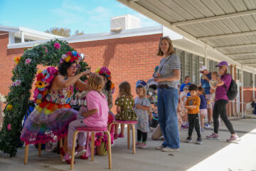 A woman in a floral dress and rainbow skirt applies glitter to a childs face at a festival booth, while children and adults wait in line nearby.