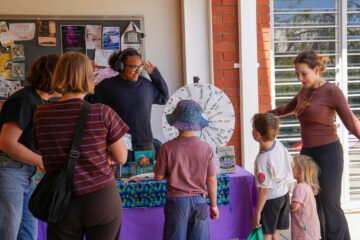 A woman wearing headphones gestures toward a prize wheel while interacting with a group of children and adults at a community table.