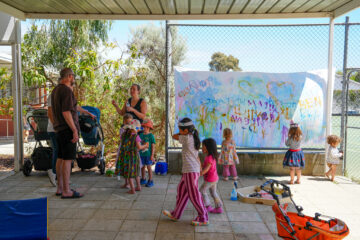 Children paint on a large paper mural attached to a chain-link fence while an adult watches nearby.