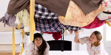 Two women in patterned shirts look up from beneath a large, suspended bundle of assorted fabrics and clothing.