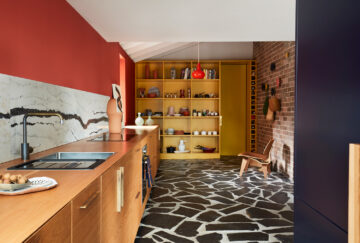 A vibrant modern kitchen with a wooden countertop, marble backsplash, and red wall. A floor-to-ceiling yellow shelving unit filled with decor stands opposite a textured brick wall with a wooden chair and black-and-white stone floor.