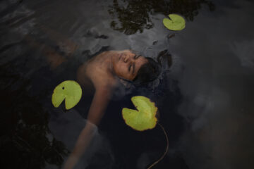A person floats on their back in dark water surrounded by green lily pads, with their eyes closed and face relaxed, partially submerged.