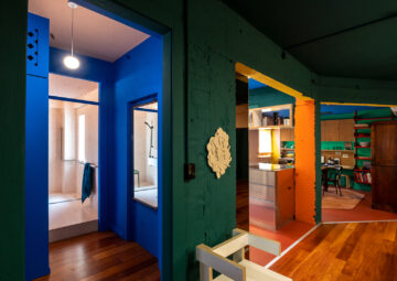 Colorful modern interior with blue, green, and yellow painted doorways, wooden floors, and a mix of contemporary and vintage furniture, including shelves and a desk in the background. Bright lighting highlights bold colors.
