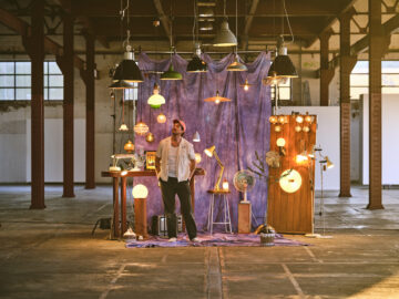 A person stands in an industrial space surrounded by various lit lamps and hanging lights, with a purple curtain backdrop and wooden panels displaying more lights, creating a cozy, artistic atmosphere.