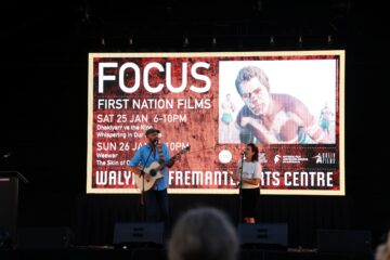 A man playing guitar and a woman speaking into a microphone stand on stage in front of a large screen displaying an event poster for Focus: First Nation Films at the Walyalup Fremantle Arts Centre.