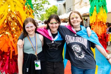 Three young people with face paint and a pride flag draped over their shoulders pose together at an outdoor event.