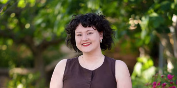 A person with short curly dark hair smiling at the camera in front of a leafy garden.