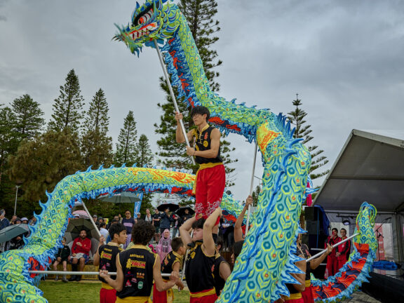 Performers in black and red uniforms hold up a long, blue and green decorative dragon during a traditional dance performance outdoors.
