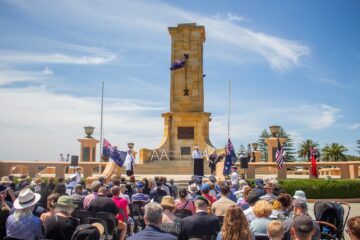 A crowd gathered at an outdoor memorial service for Anzac Day, with speakers standing at a podium before a large stone monument and flags at half-mast.