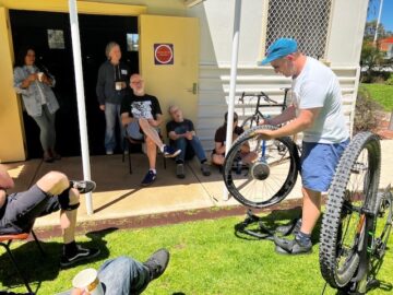 A man in a blue cap demonstrates how to repair a bicycle tire to a small group of people sitting outside a building.