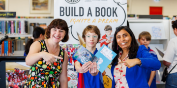Two women and a boy hold up books titled Fin the Fish Wizard in front of a Build a Book event sign in a library.