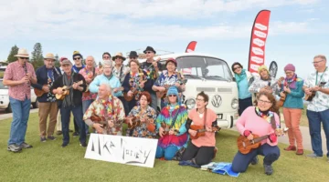 A large group of ukulele players posing in front of a white Volkswagen van on a grassy lawn, with one member holding a sign that reads Uke Society.