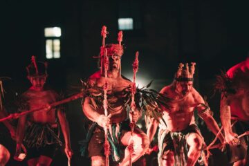 Several people in traditional ceremonial attire and headpieces perform a dance while holding wooden staves under red lighting.