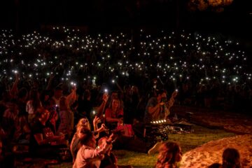 A large crowd of people sitting on the grass at night, holding up their illuminated smartphones.