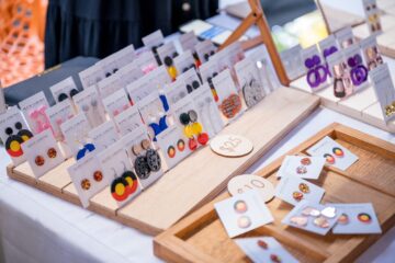 Display of various Buffie Coruna brand earrings on wooden stands, including round designs featuring Aboriginal flag colors and other decorative shapes, with price tags of $10 and $25.