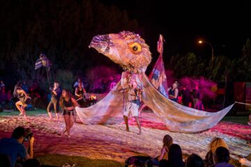 Performers in a nighttime outdoor show dance around a person wearing a large, textured bird costume with expansive wings.