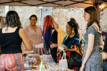 A woman at a craft market holds up a red and blue scarf for shoppers to see.