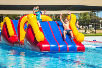 Two boys slide down a large, red, yellow, and blue inflatable water slide into a swimming pool.