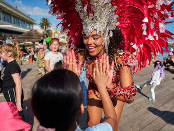A performer in a red feathered carnival costume and rhinestone headdress high-fives a child on a wooden boardwalk.