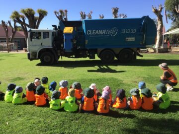 A group of children in high-visibility vests sit on the grass watching a parked blue Cleanaway garbage truck.