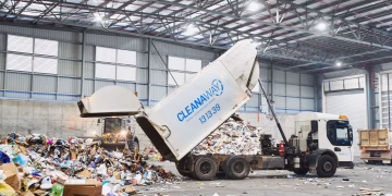 A Cleanaway waste collection truck lifts its container to dump a load of recyclables in a large warehouse facility.