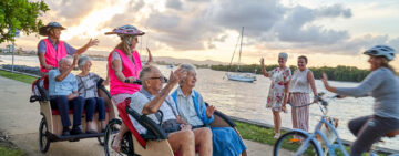 A group of people enjoying a sunny day by the water, with some riding in rickshaws and others biking or waving.