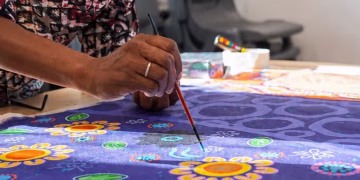 A person paints colorful floral and abstract patterns on a purple fabric, using a thin brush. The artwork includes yellow flowers, blue swirls, and green leaves.