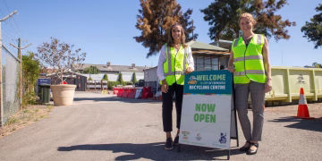 Two women in high-visibility vests stand next to a sign for the City of Fremantle Recycling Centre that reads Welcome and Now Open.