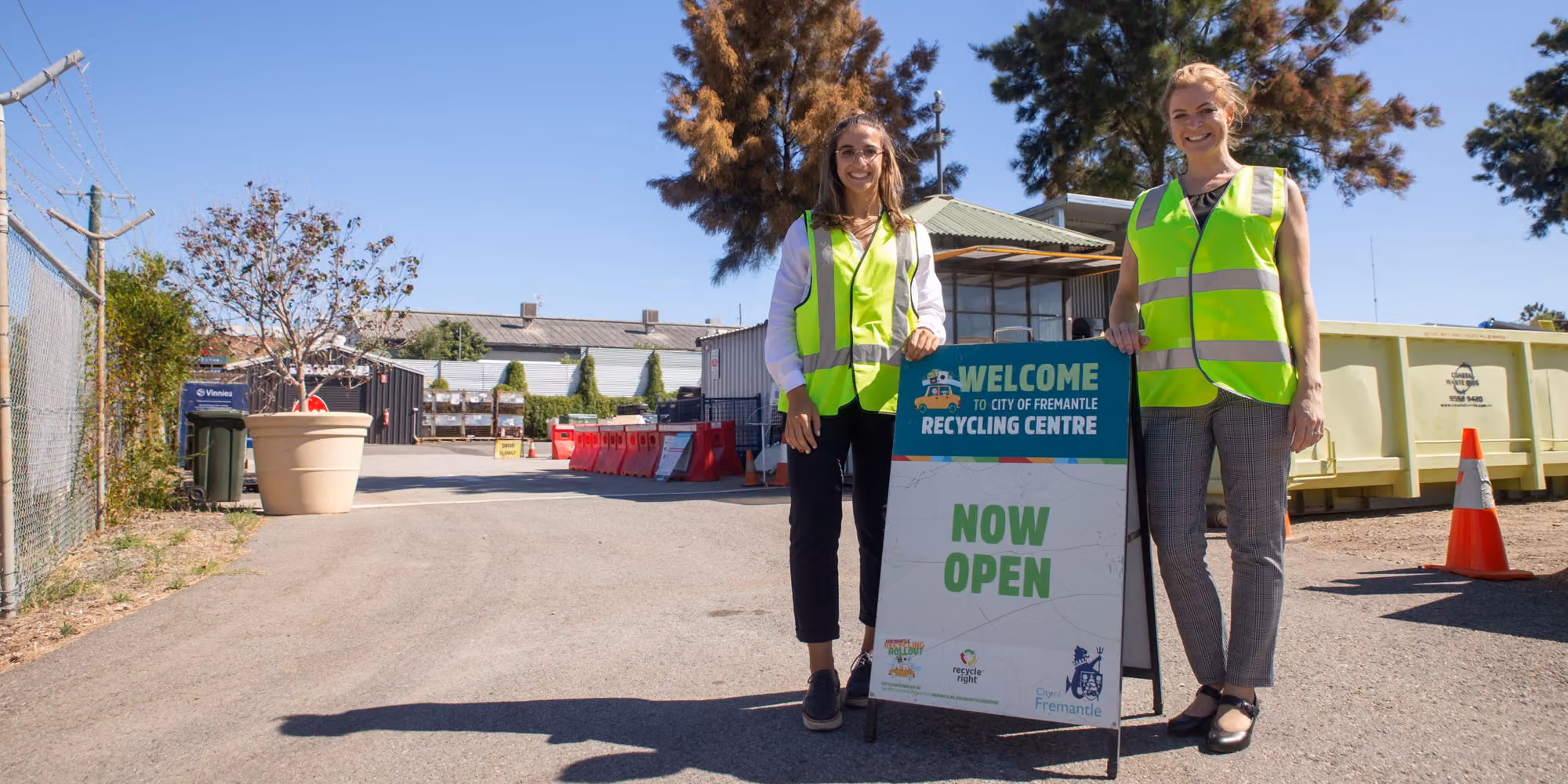 Two women in high-visibility vests stand next to a sign for the City of Fremantle Recycling Centre that reads Welcome and Now Open.